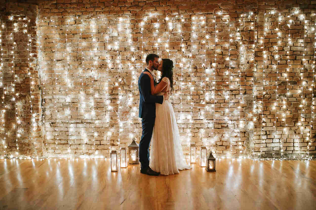 Casal de noivos se beijando em frente à uma parede coberta por fairy lights