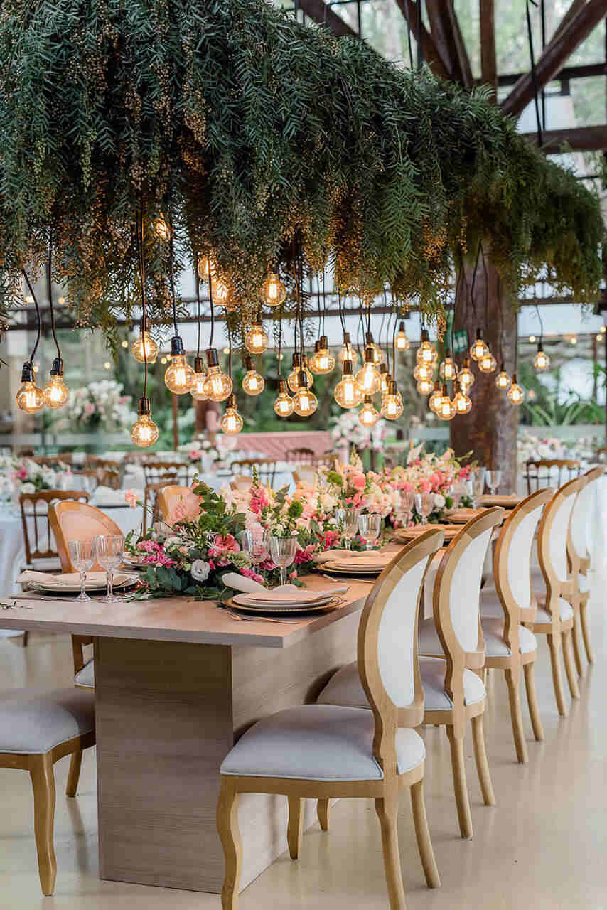 Mesa dos convidados com flores, pratos e talheres. Acima deles há folhagens e luzes penduradas