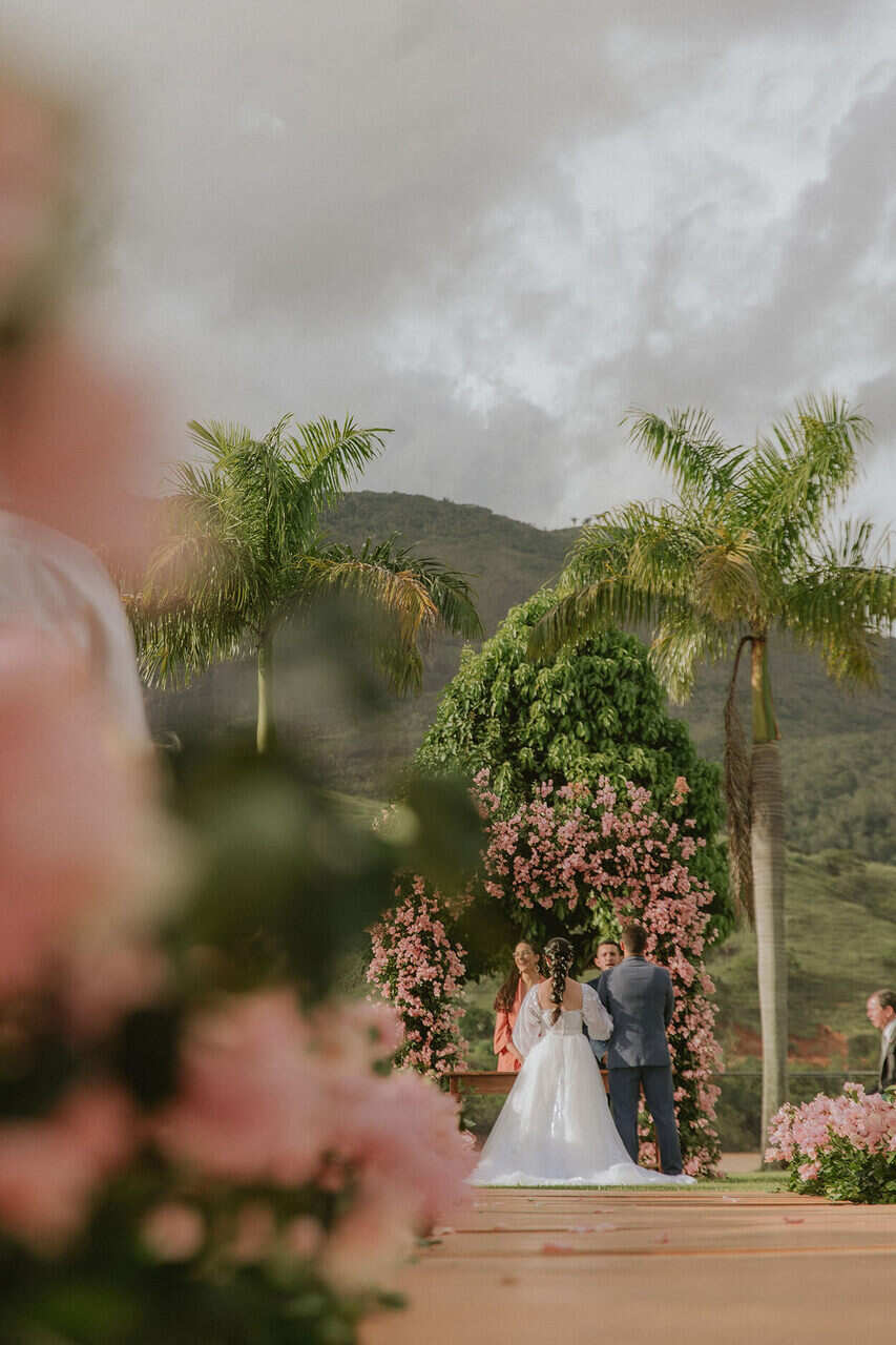 Casamento ao ar livre. Os noivos no altar de costas para os convidados. Em frente ao casal, há um arco de flores rosas.