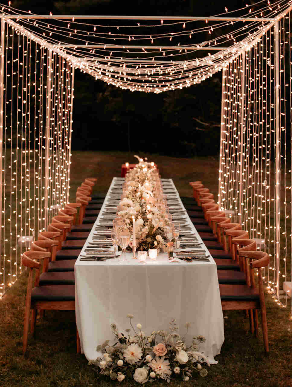 Mesa dos convidados, com as cadeiras devidamente alinhadas. Arranjos florais e vela sob a mesa e um varal de luzes acima, cobrindo toda a mesa