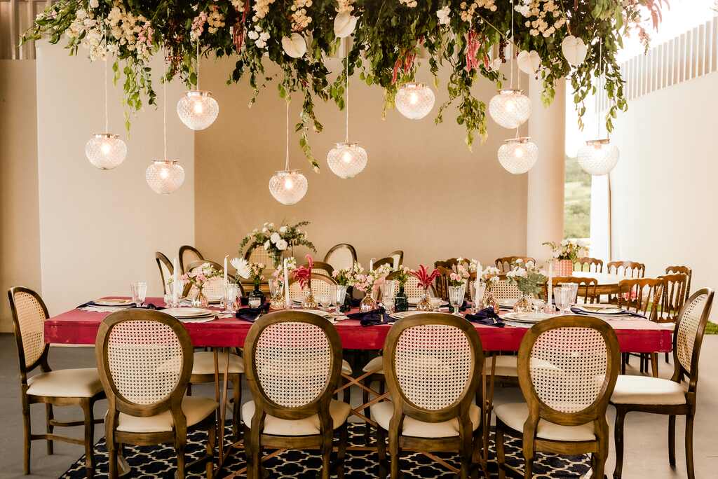 Mesa dos convidados com as cadeiras vintage, velas, louças e flores sob a mesa. Acima dela, flores, folhagens e pendentes de vidro pendurados.