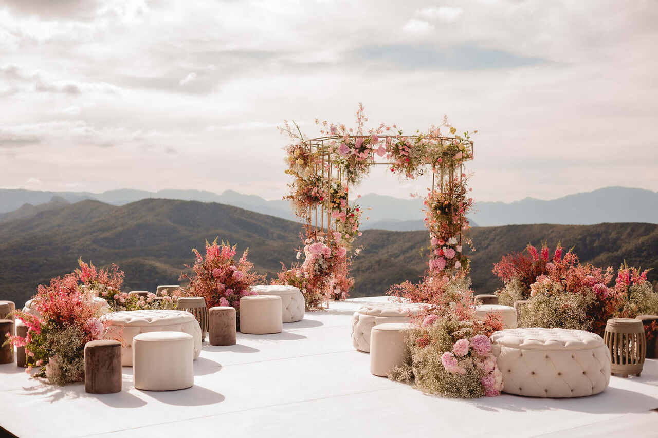 Cenário de casamento no topo de uma montanha, com puffs claros, vasos de flores entre eles e um arco de flores no altar.
