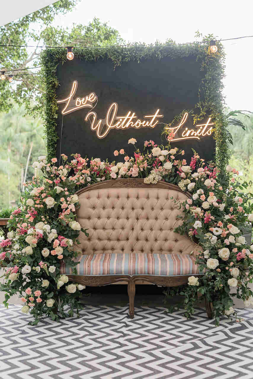 Sofá vintage rodeado de flores. Ao fundo, um painel com um letreiro em neon pendurado