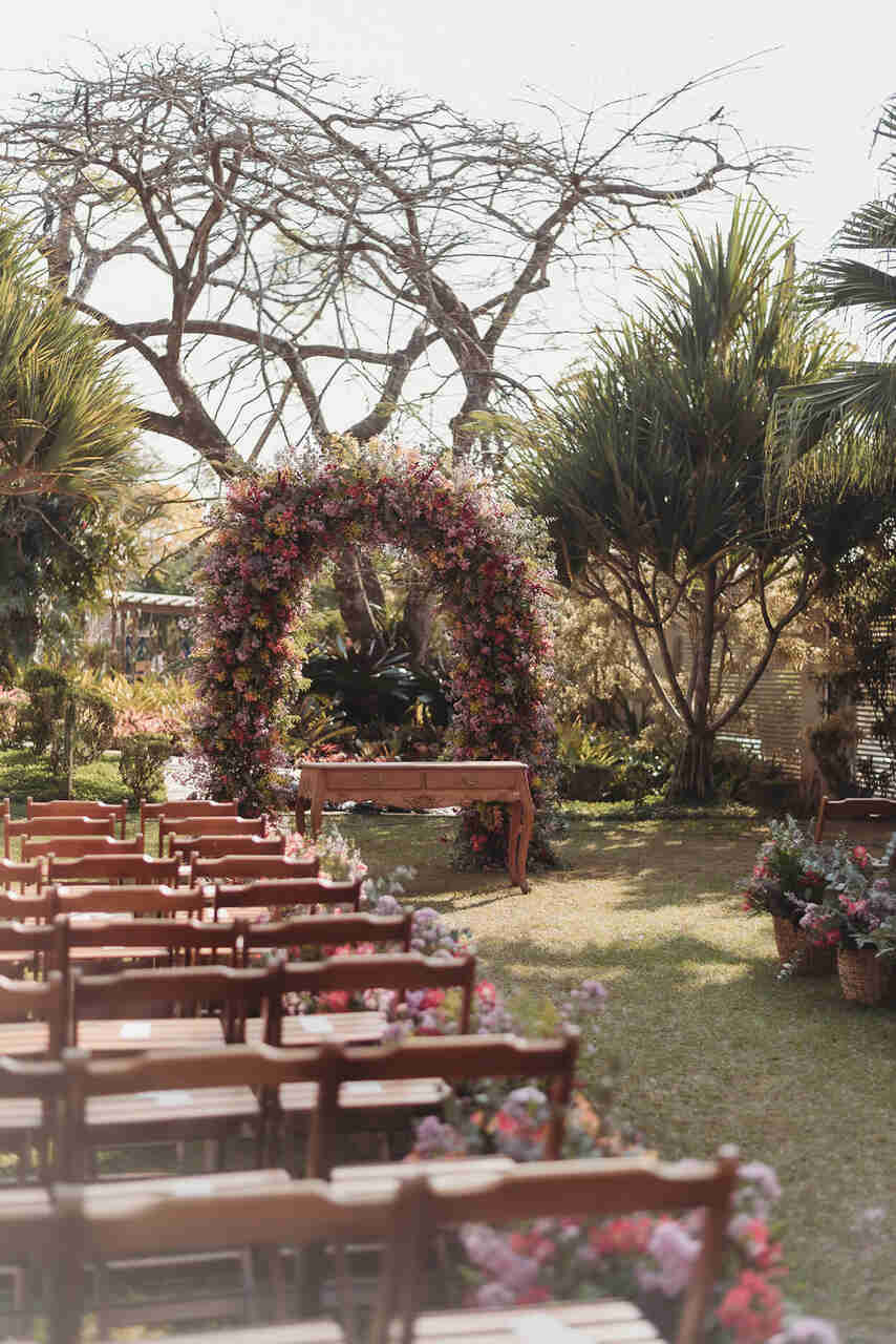 Cenário de um casamento ao ar livre, com bancos de madeira. No altar, há um aparador de madeira e um arco de flores.