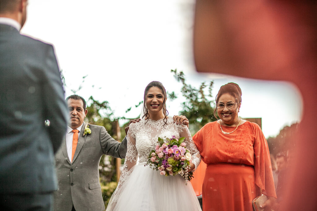 entrada da noiva com a mãe co. vestido laranja de manga longa e pai com terno cinza e gravata laranja
