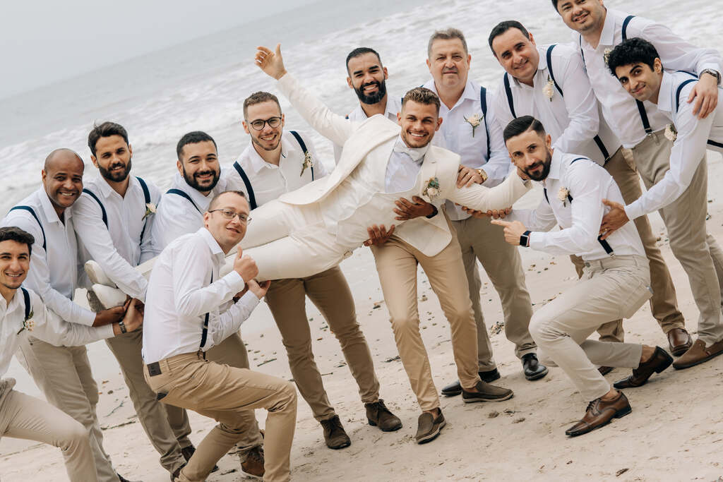 padrinhos com calça caqui segurando camisa branca com suspensório preto carregando o noivo na praia