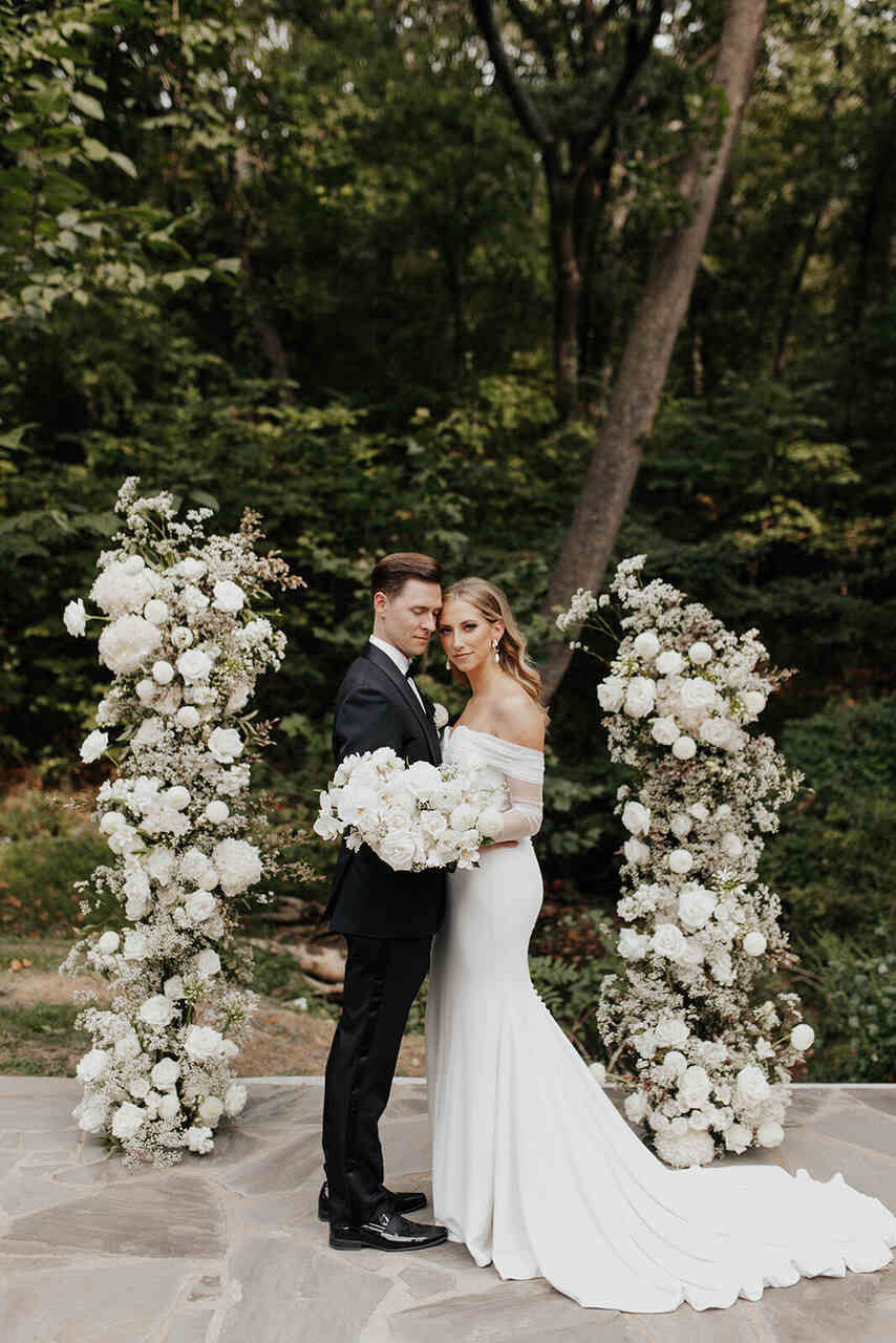 Casal posando para a foto. Noiva segurando um buquê e olhando para a foto e o noivo com o rosto encostado na noiva, olhando para ela. Ao lado deles há pilares pequenos com flores brancas