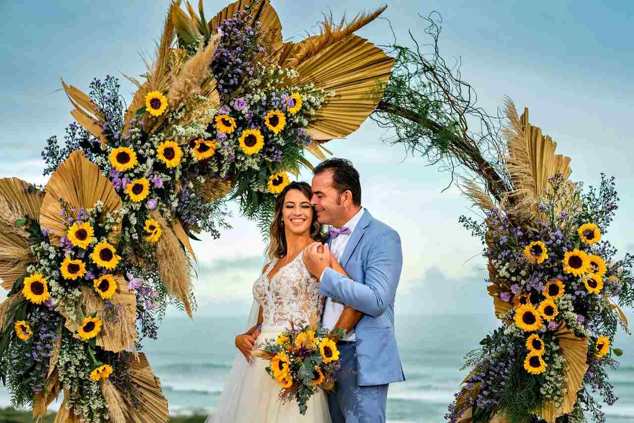 Noivos no altar com um arco de flores feito de girassóis, folhagens e gipsofilas, com o mar ao fundo