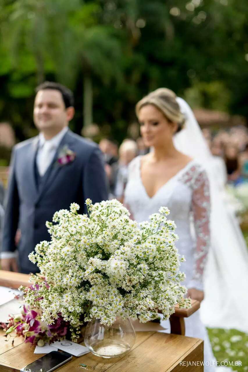 Arranjo de áster em cima da aparado do altar. Ao fundo, desfocado, o casal de noivos