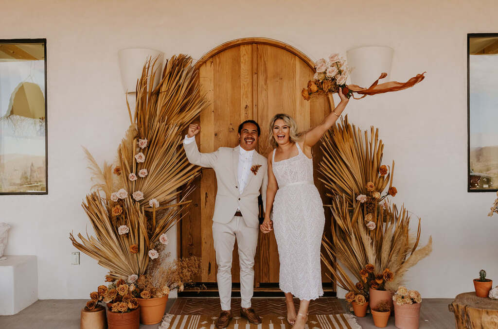 Casal posando para a foto levantando os braços. Em volta deles há uma decoração com folhagens e flores secas