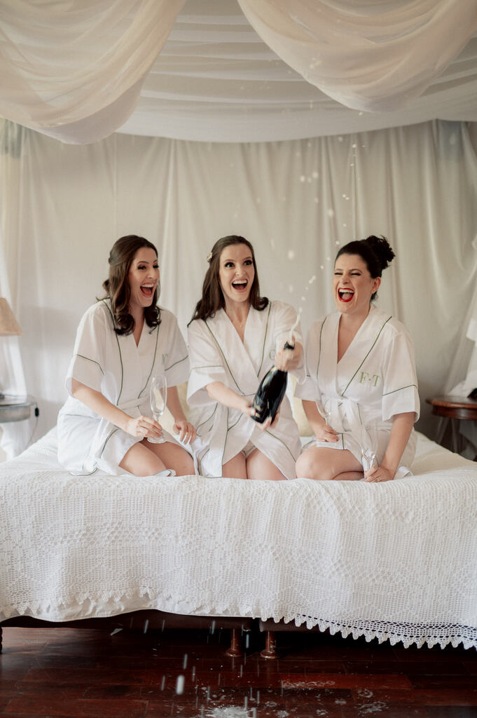 Noiva abrindo um espumante com duas convidadas especiais. As três estão sentadas na cama