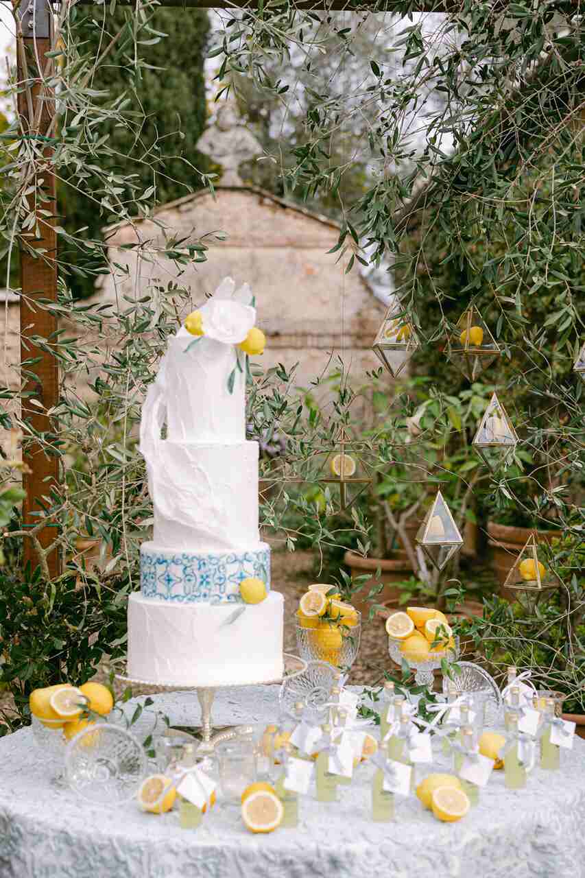 Bolo de 3 andares branco no topo com uma flor branca de limões sicilianos em cima da mesa com cristaleiras e limões