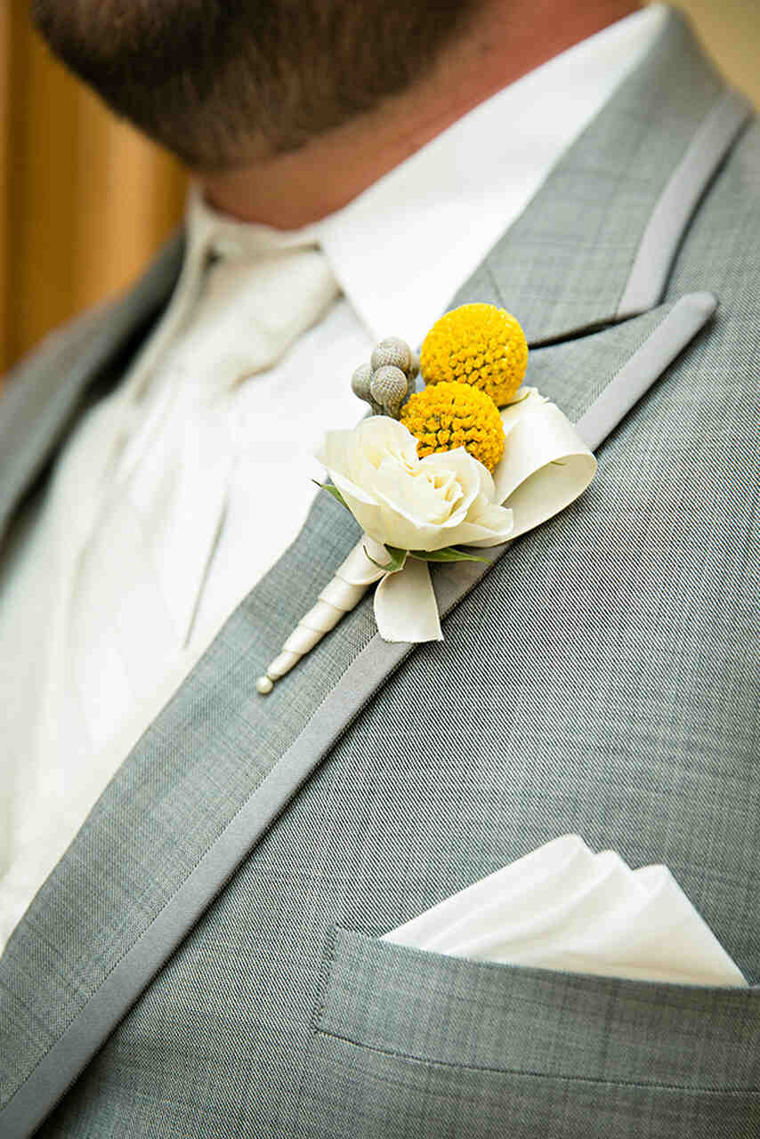 Detalhes do traje do noivo, num terno cinza claro com botonniere de flor amarela