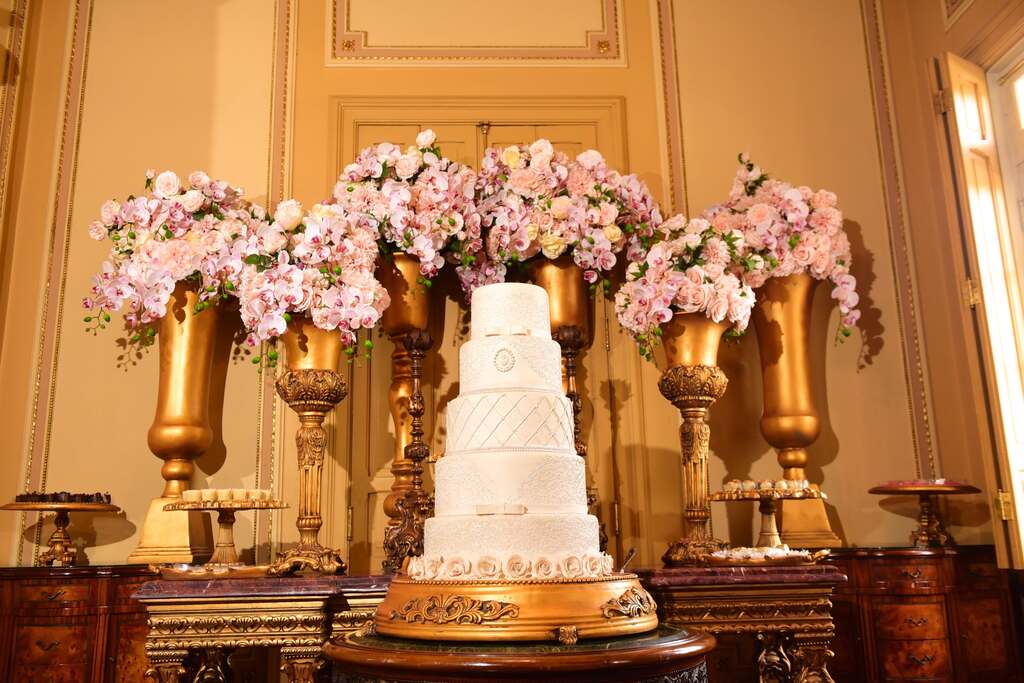 bolo de casamento fake com 5 andares e ao fundo vasos dourados com flores cor de rosa