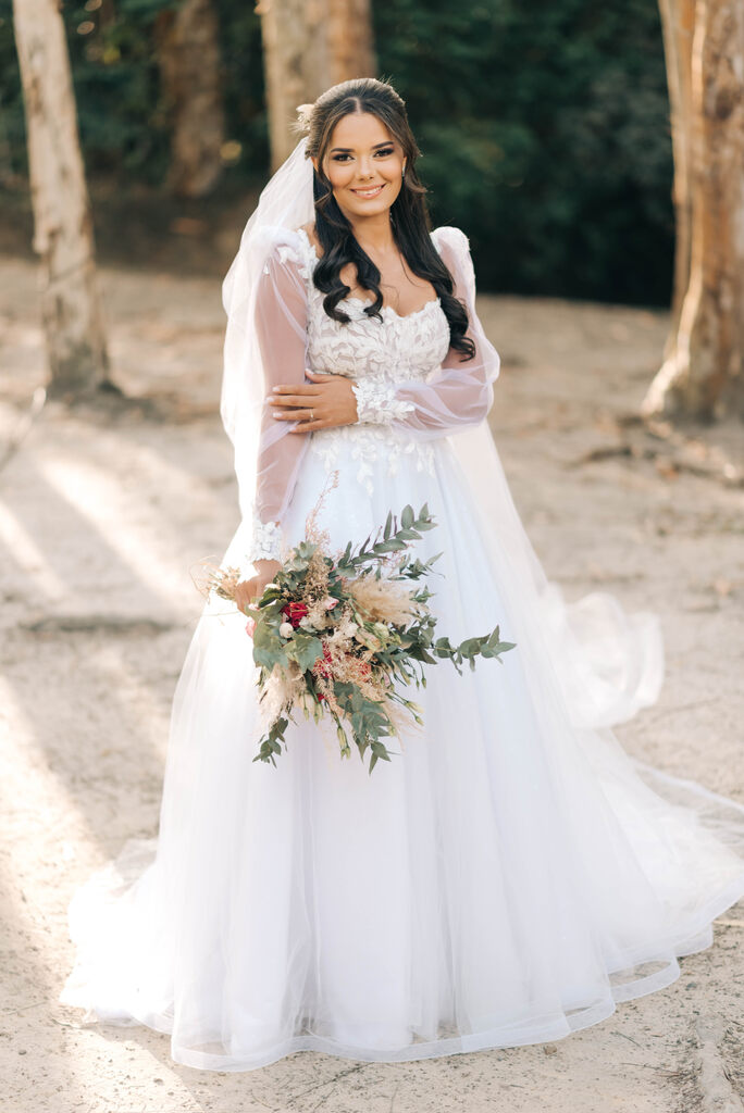 mulher com vestido de noiva com manga longa segurando buquê e com cabelo semi preso e véu