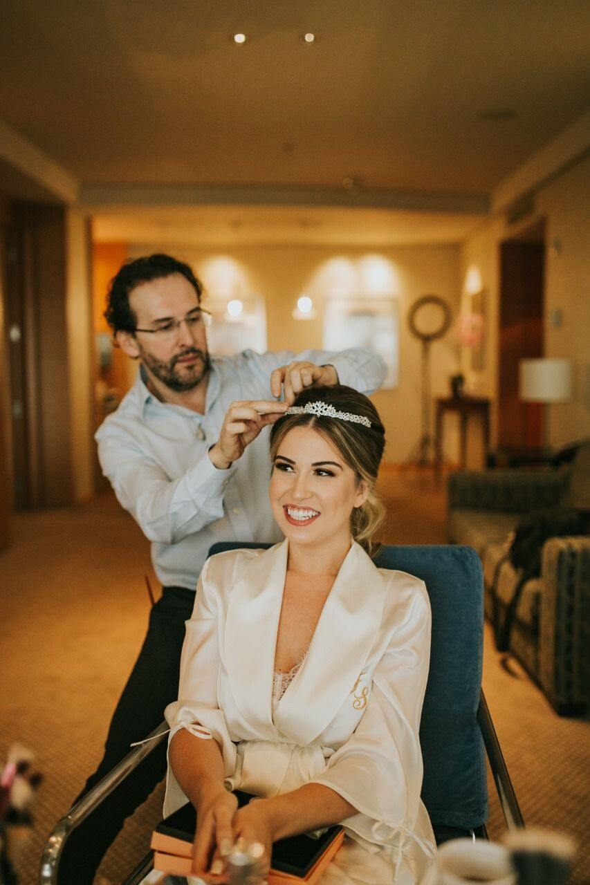 profissional dando os últimos retoques no penteado da noiva. A noiva está sentada sorrindo