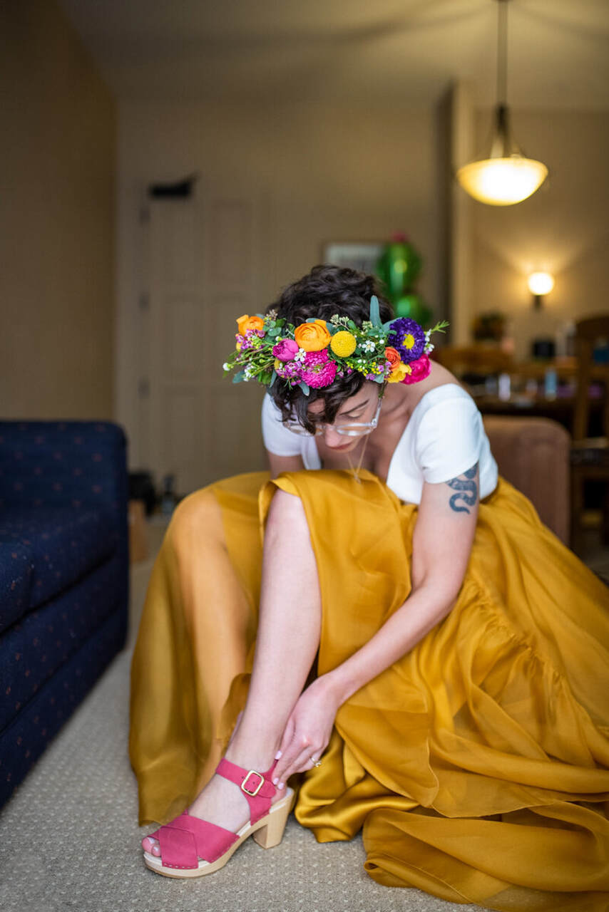 Noiva ajeitando o sapato rosa. Ela está com uma saia mostarda, blusa branca e coroa de flores coloridas na cabeça