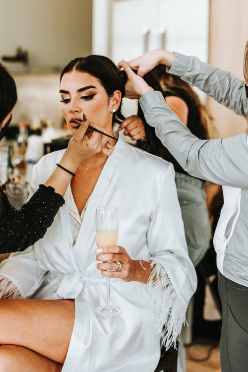 Noiva sentada com uma taça de bebida na mão. Uma profissional passando batom na boca da noiva enquanto outra mexe no cabelo dela para fazer o penteado