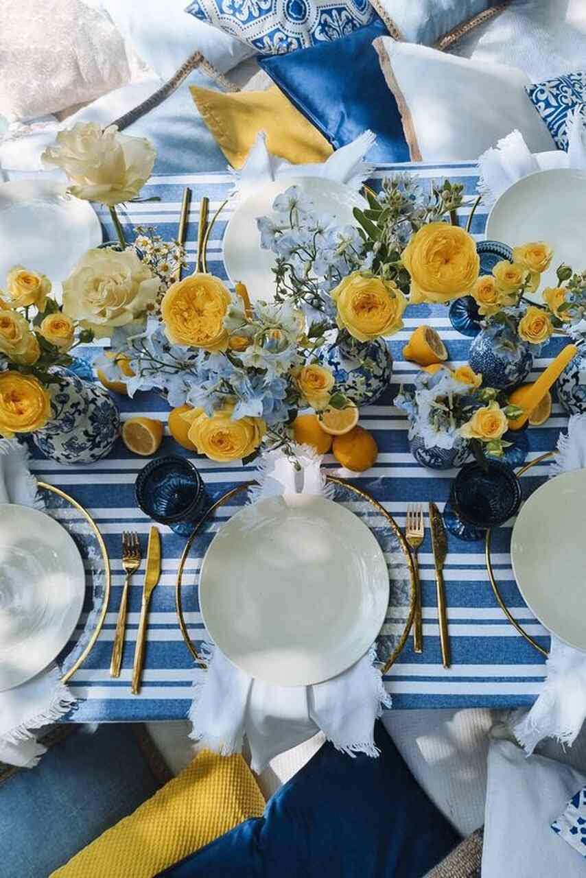 Mesa dos convidados com toalha listrada azul e branco, pratos brancos e arranjos florais amarelos