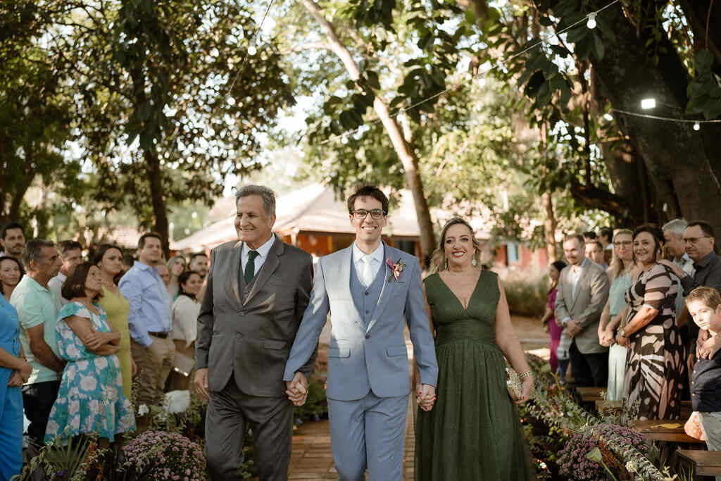 entrada do noivo com terno azul claro ao lado do pai com terno cinza e mãe com vestido verde escuro