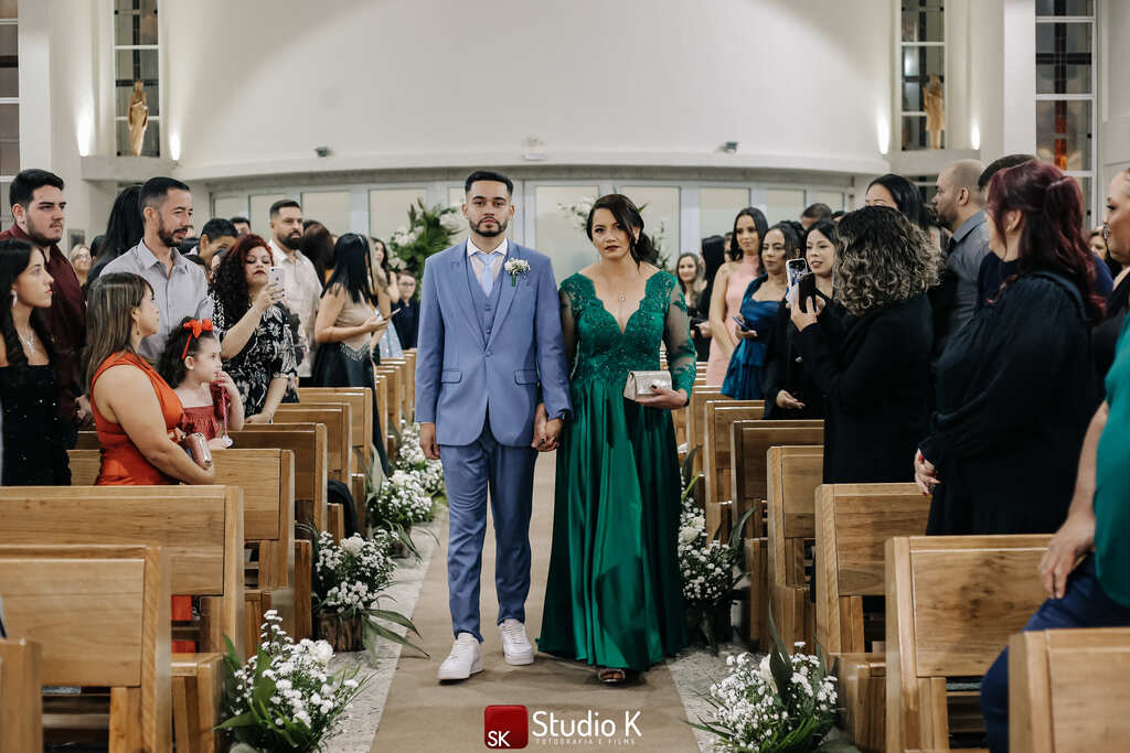 entrada do noivo com tenro azul com colete ao lado da mãe com vestido verde esmeralda