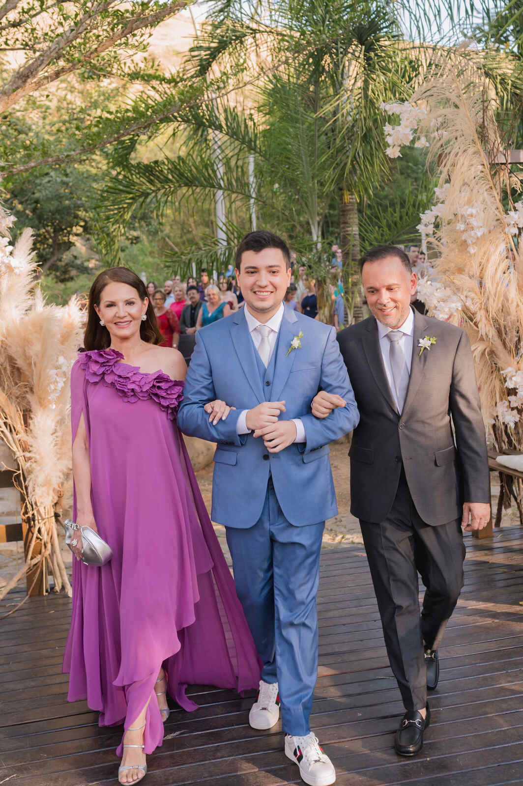 entrada do noivo com terno azul e mãe com vestido lilás e pai com terno preto