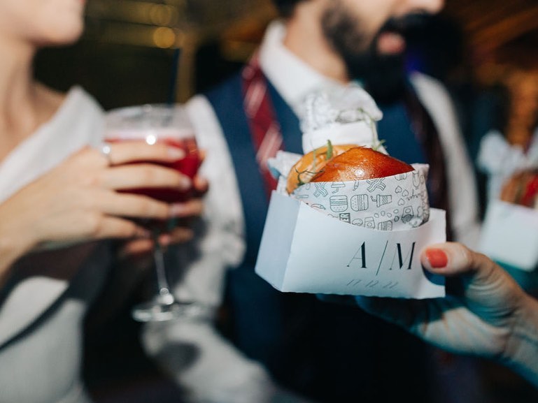 Noivo e noiva brindando com lanches personalizados durante a festa de casamento, segurando um hambúrguer e uma bebida