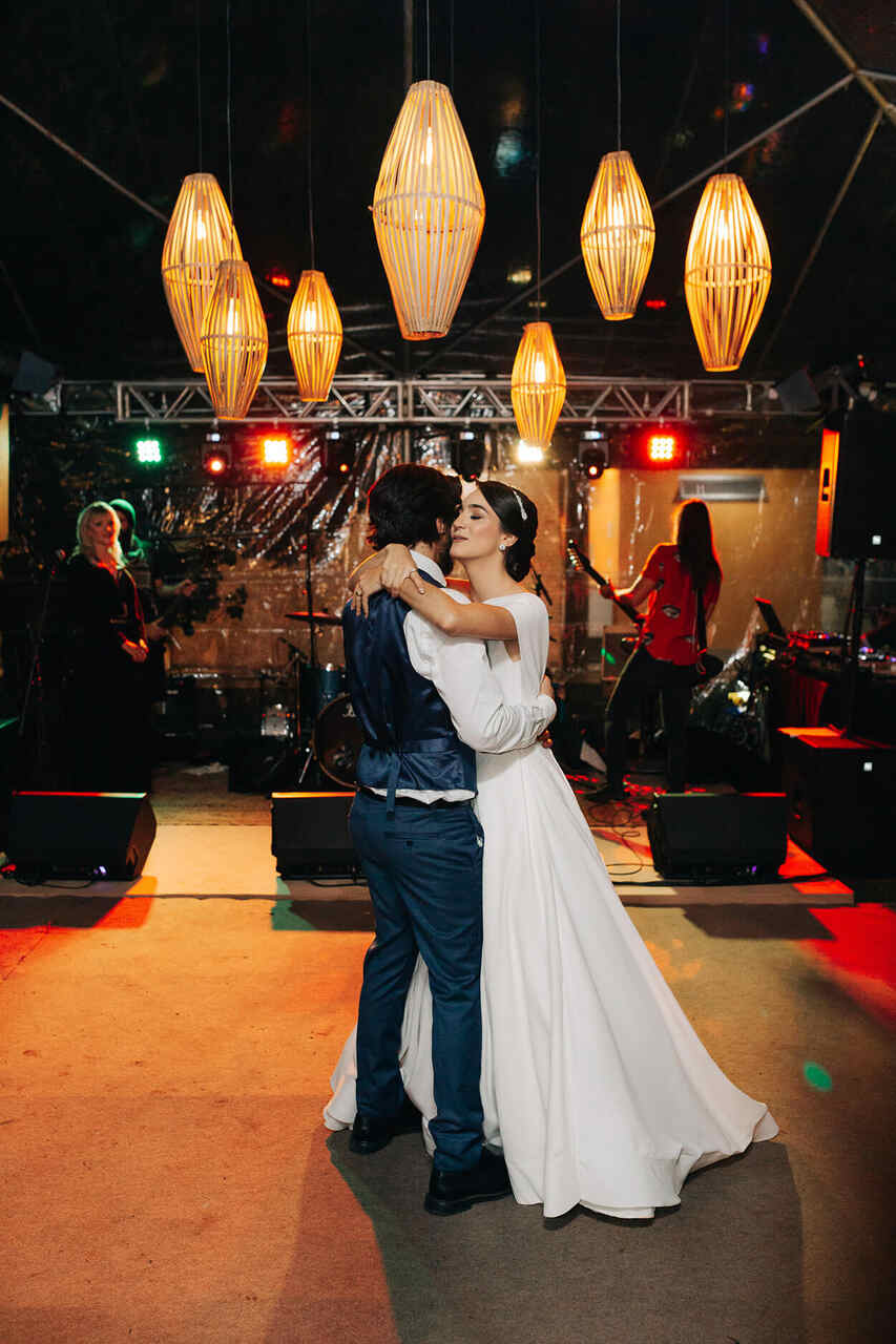 Casal de noivos dançando juntos em um espaço decorado com luminárias pendentes, em um ambiente de casamento com banda ao fundo