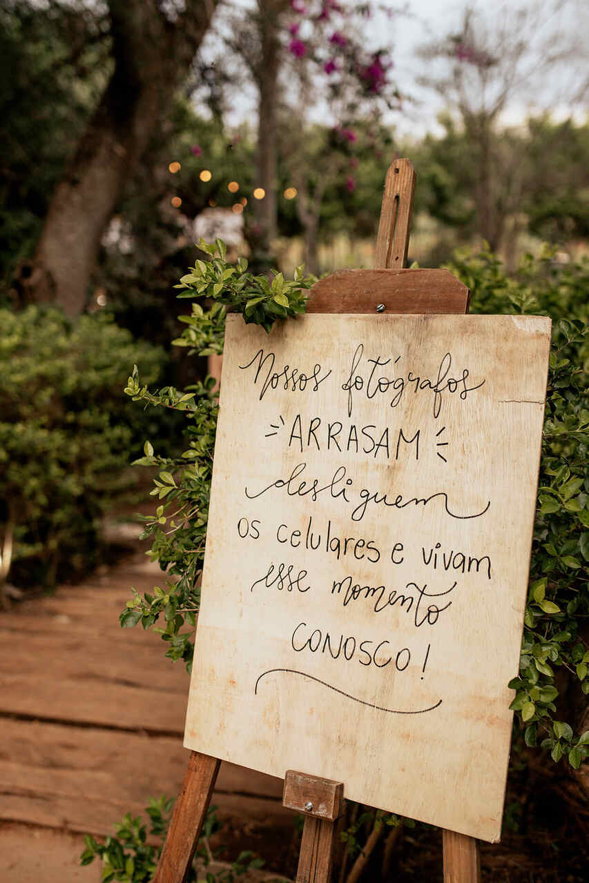 Placa de madeira em cima de um cavalete escrito: Nossos fotógrafos arrasam. Desliguem os celulares e vivam esse momento conosco.