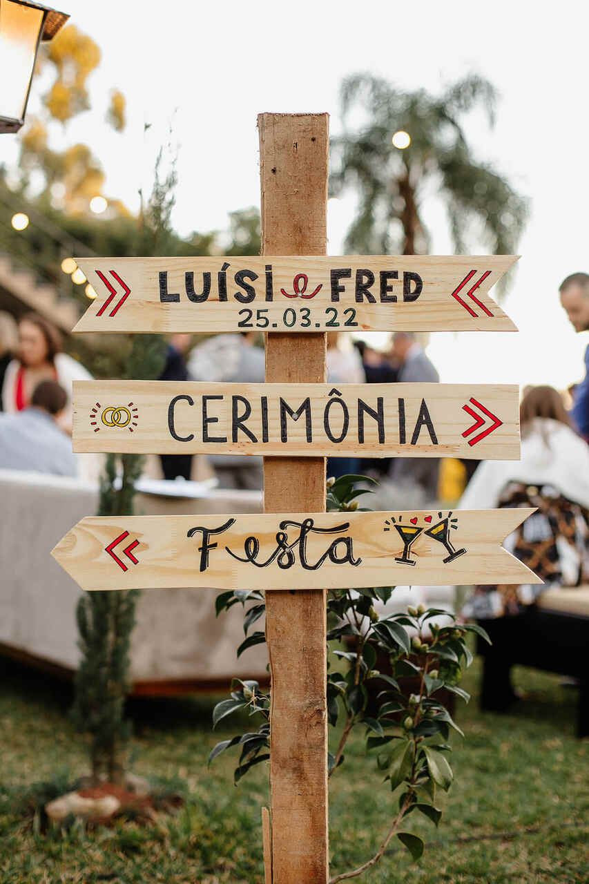 Placa fincada no chão, dividida em três partes, indicando onde é a cerimônia e a festa