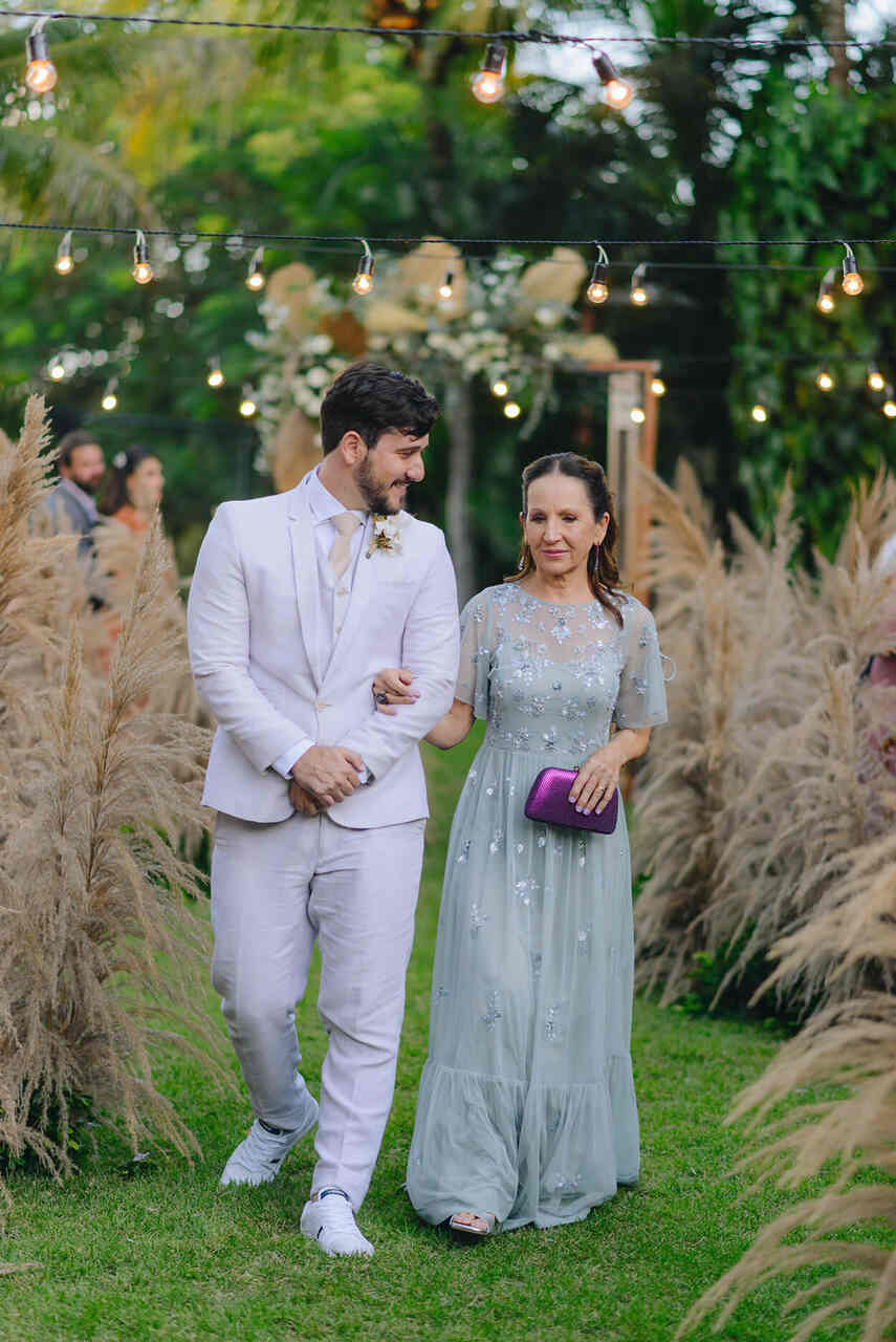 Noivo entrando na cerimônia com a sua mãe que está de vestido em tom pastel e uma bolsa de mão roxa