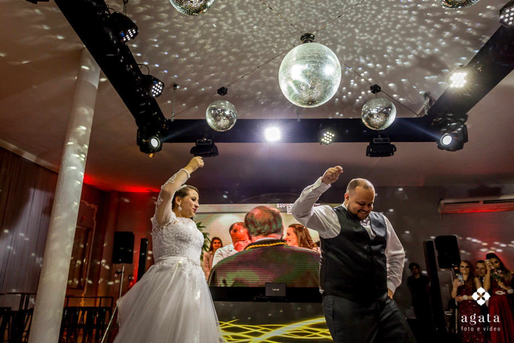 Casal de noivos sorrindo e dançando alegremente sob um globo espelhado em sua festa de casamento