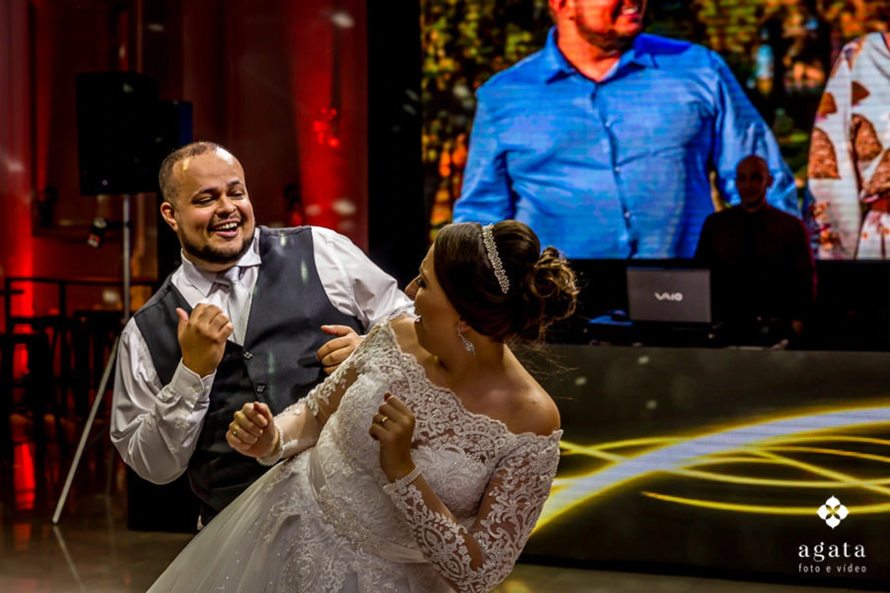 Casal de noivos sorrindo e dançando animadamente durante a festa de casamento. A noiva, com um vestido rendado e tiara, olha para o noivo, enquanto ele, vestindo de colete e gravata, retribui o olhar 