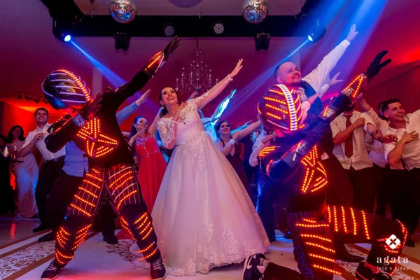 capa post: Noiva e noivo celebrando com dançarinos em trajes de LED durante a festa de casamento. A noiva, sorridente e com vestido de renda, faz um gesto animado ao lado do noivo e dos dançarinos, que estão vestidos com trajes futuristas iluminados por luzes vermelhas