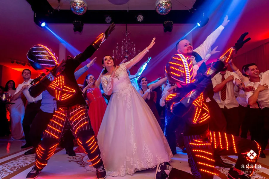 Noiva e noivo celebrando com dançarinos em trajes de LED durante a festa de casamento. A noiva, sorridente e com vestido de renda, faz um gesto animado ao lado do noivo e dos dançarinos, que estão vestidos com trajes futuristas iluminados por luzes vermelhas