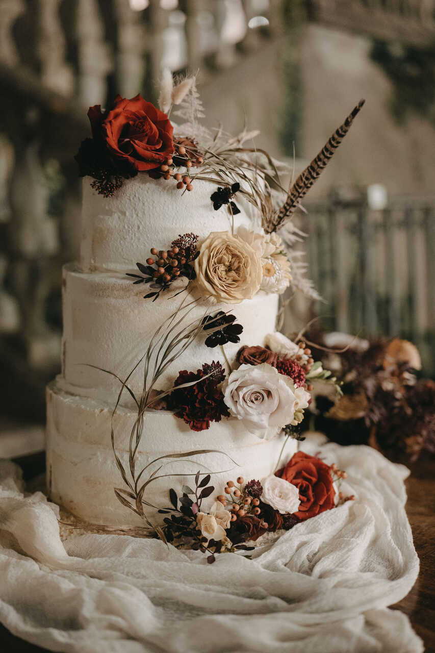 Bolo de casamento rústico de três camadas, decorado com flores naturais em tons de vermelho e creme, folhas secas e detalhes delicados