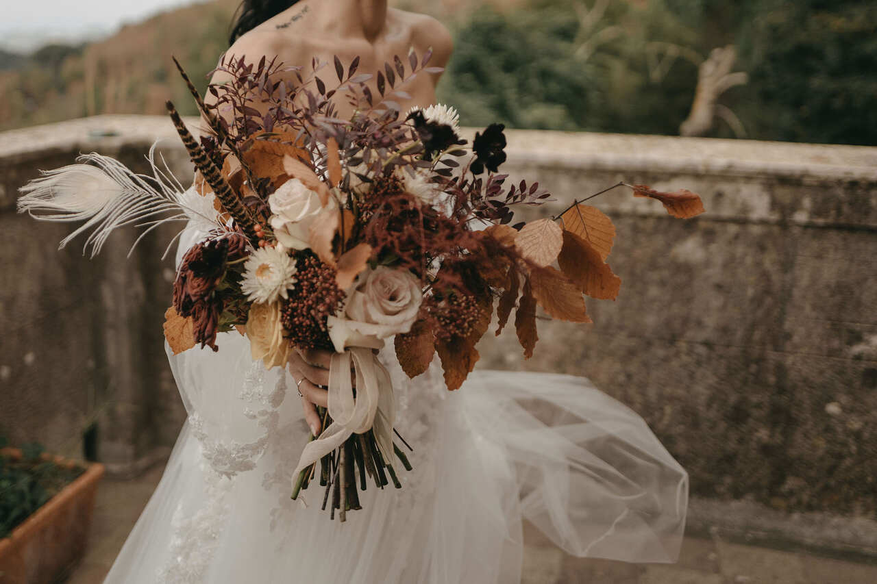 Buquê de noiva em tons outonais, com flores secas, folhas douradas, rosas brancas e detalhes de penas