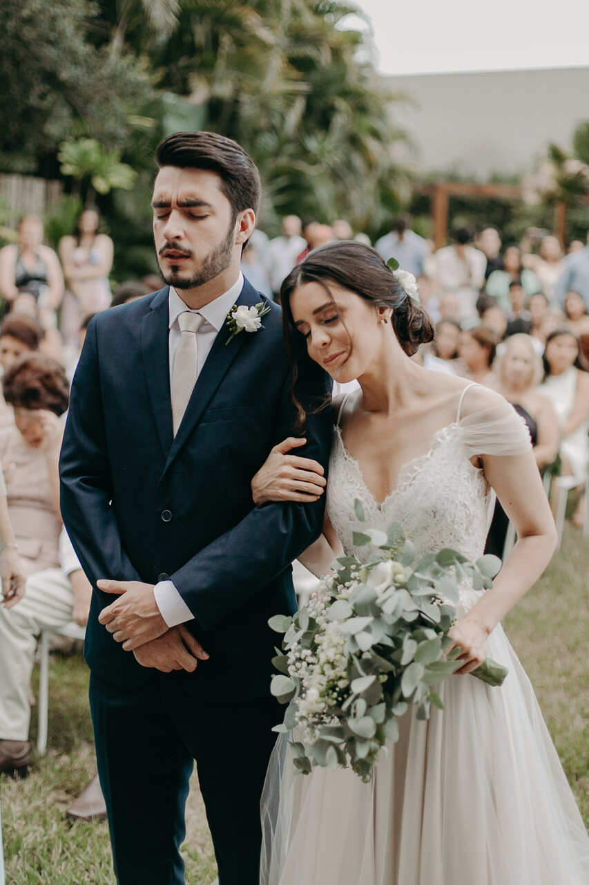 Casal de olhos fechados no altar. Noiva encosta cabeça no ombro do noivo