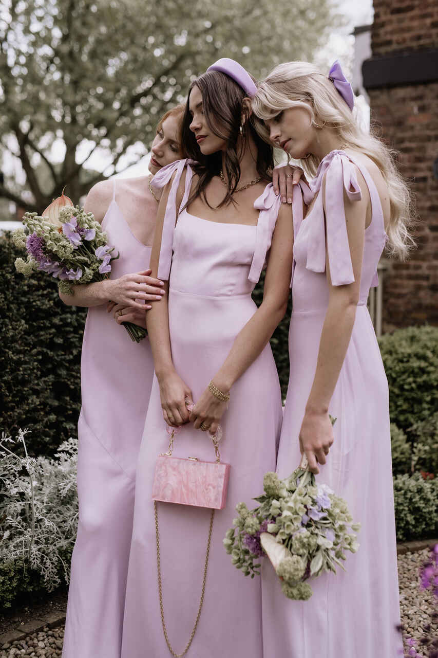 Três damas com vestido rosa claro. As duas da ponta seguram buquês e a que está ao centro, segura uma bolsa da mesma cor do vestido