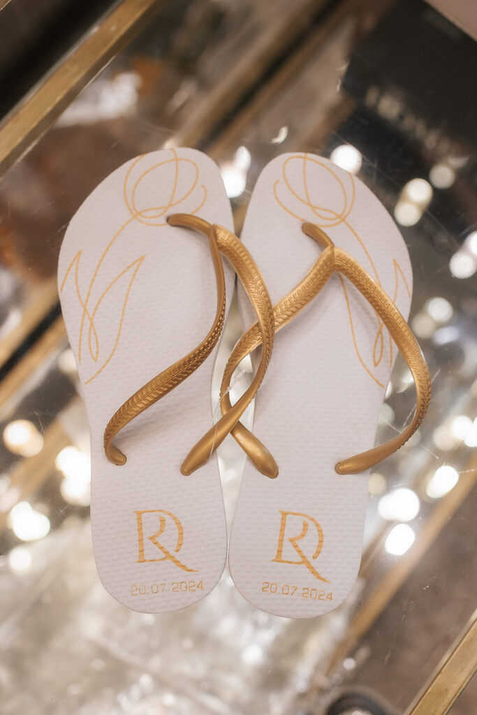 Chinelo personalizado branco com detalhes dourados, incluindo as iniciais dos noivos e a data do casamento, 20.07.2024, sobre uma superfície espelhada