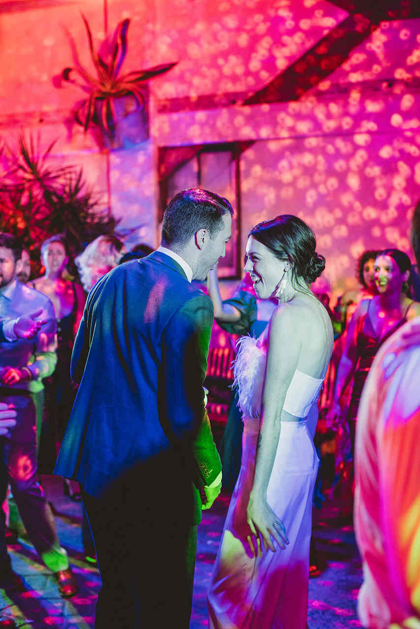 Casal de noivos sorrindo e se divertindo na pista de dança, cercados por convidados e uma iluminação colorida vibrante