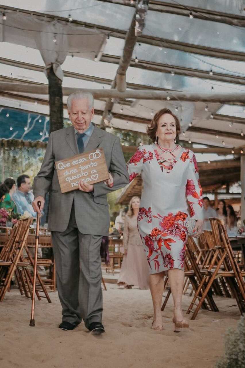 Casal de avós entrando na cerimônia. O homem segura uma placa escrita Que essa aliança dure tanto quanto a nossa