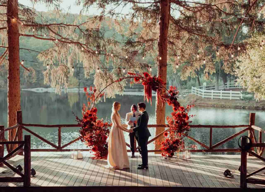 Casal no altar de frente um para o outro. O cenário da cerimônia é em um deck, com um lago ao fundo e no altar há um arco de folhas e flores em cor terrosos