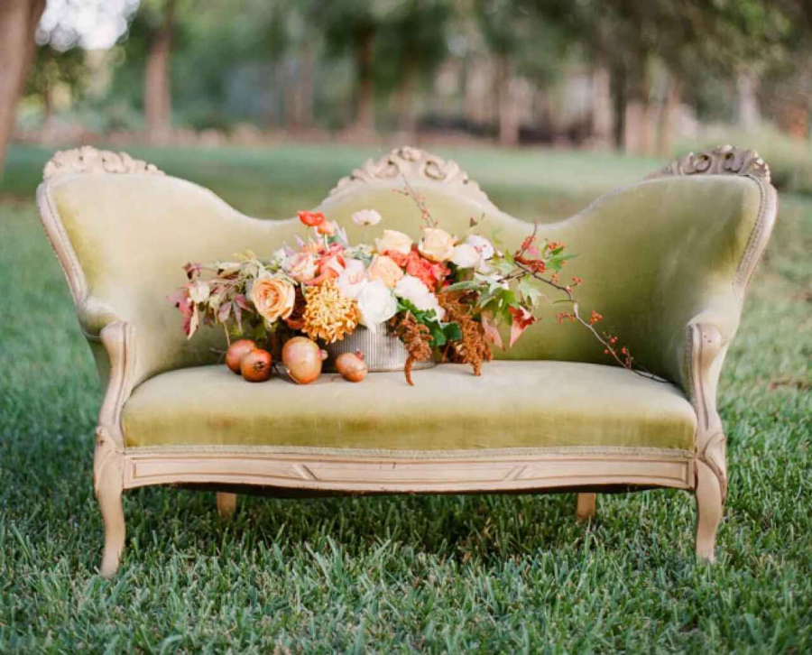 Sofá retrô na cor verde e ao centro um buquê com flores, folhas e frutas