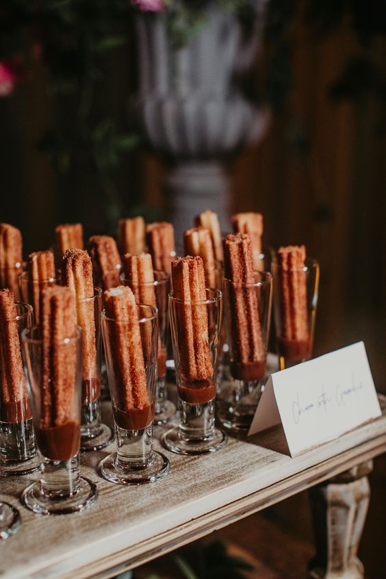 Bandejas de churros servidos em copos de vidro com chocolate derretido no fundo. Cada copo contém um churro em pé.