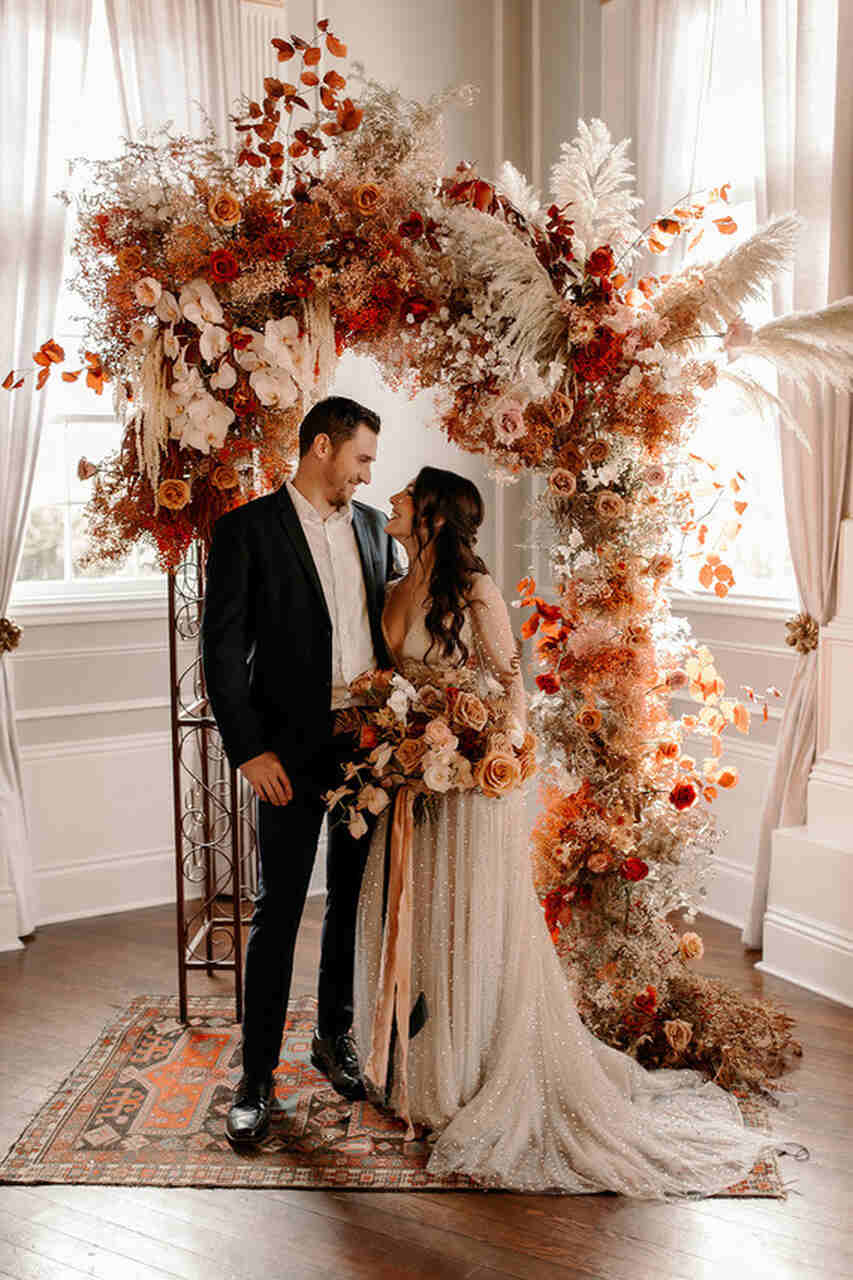 Casal em frente ao arco de flores e folhas, de mãos dadas e olhando um para o outro