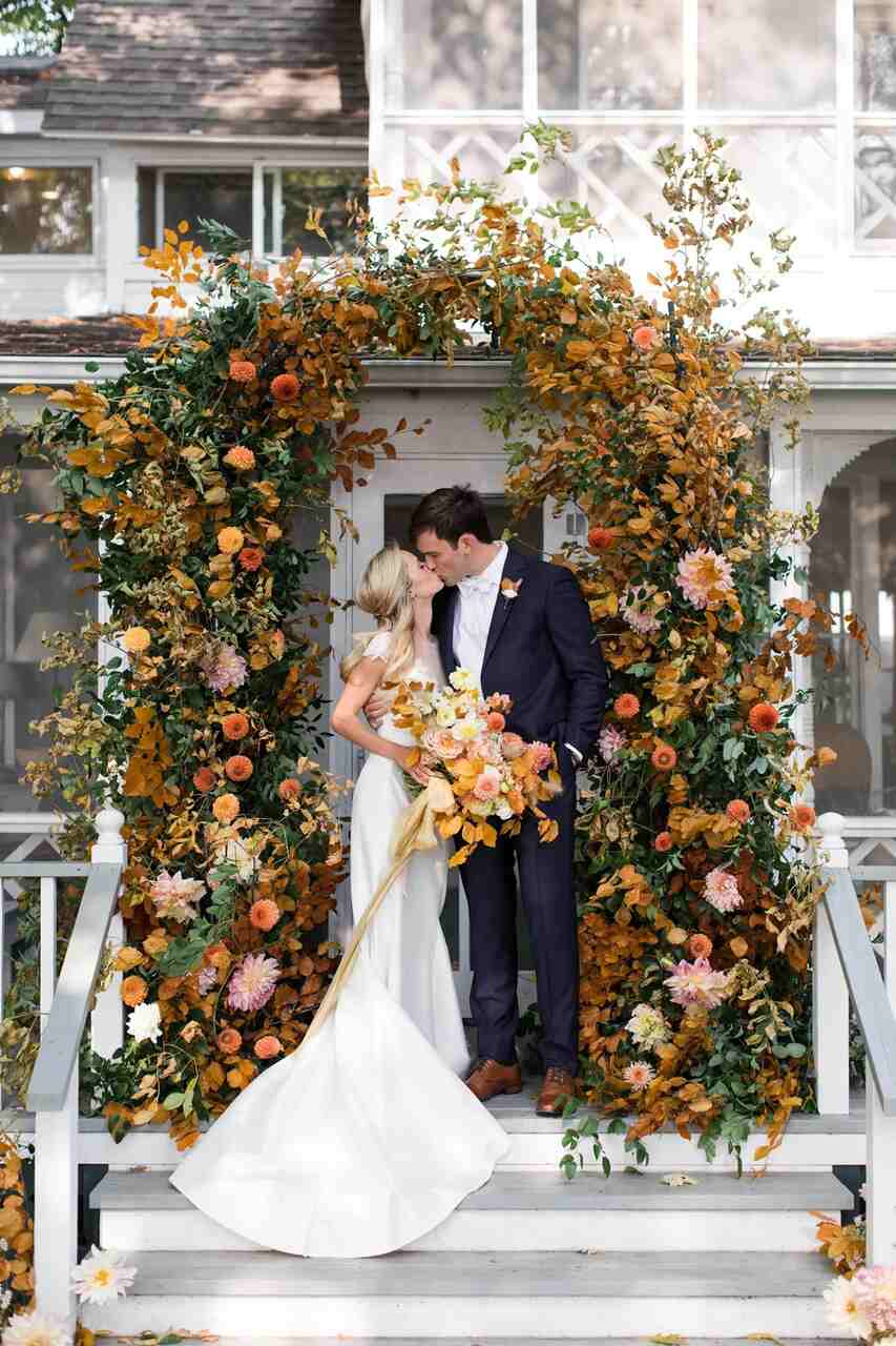 Casal se beijando dentro de um arco de flores e folhas em tons terrosos