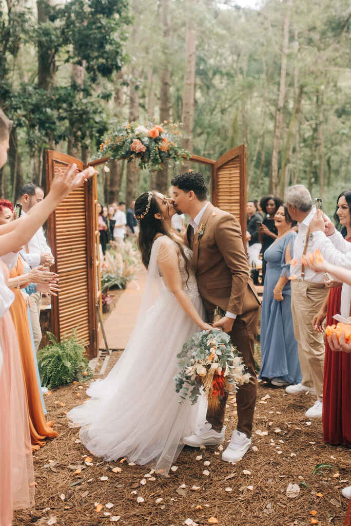 Noivos se beijam na saída da cerimônia ao ar livre, com convidados celebrando ao redor