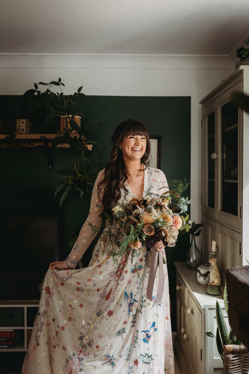 Noiva sorrindo, com uma das mãos segurando o vestido que é com rendas coloridas em flor e a outra mão segurando o buquê