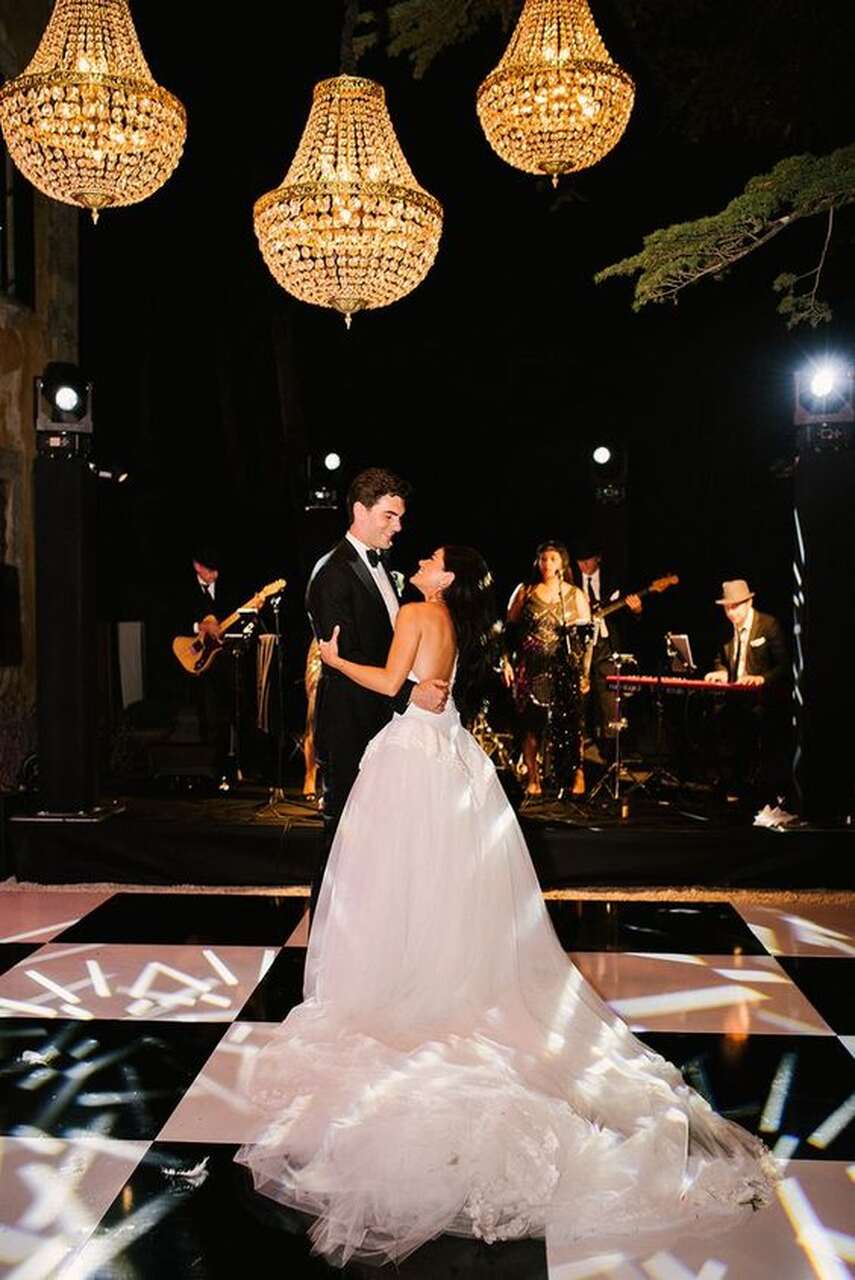 Noivos dançando em uma pista iluminada em preto e branco, sob lustres grandes e luxuosos, enquanto uma banda ao fundo toca